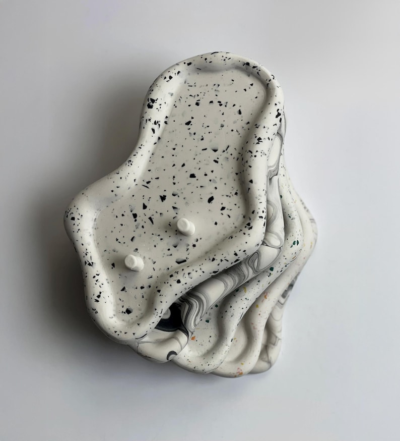 May include: A stack of four irregularly shaped, speckled white trays with black flecks. The top tray has two small, white, circular protrusions. The edges of the trays have a wavy design, and the bottom trays have a marbled pattern.