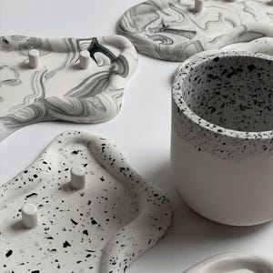 May include: An assortment of decorative trays and a small cup. The trays feature unique designs, including a marbled pattern in shades of gray and white, and a speckled terrazzo-like design. The cup is white with a speckled rim.