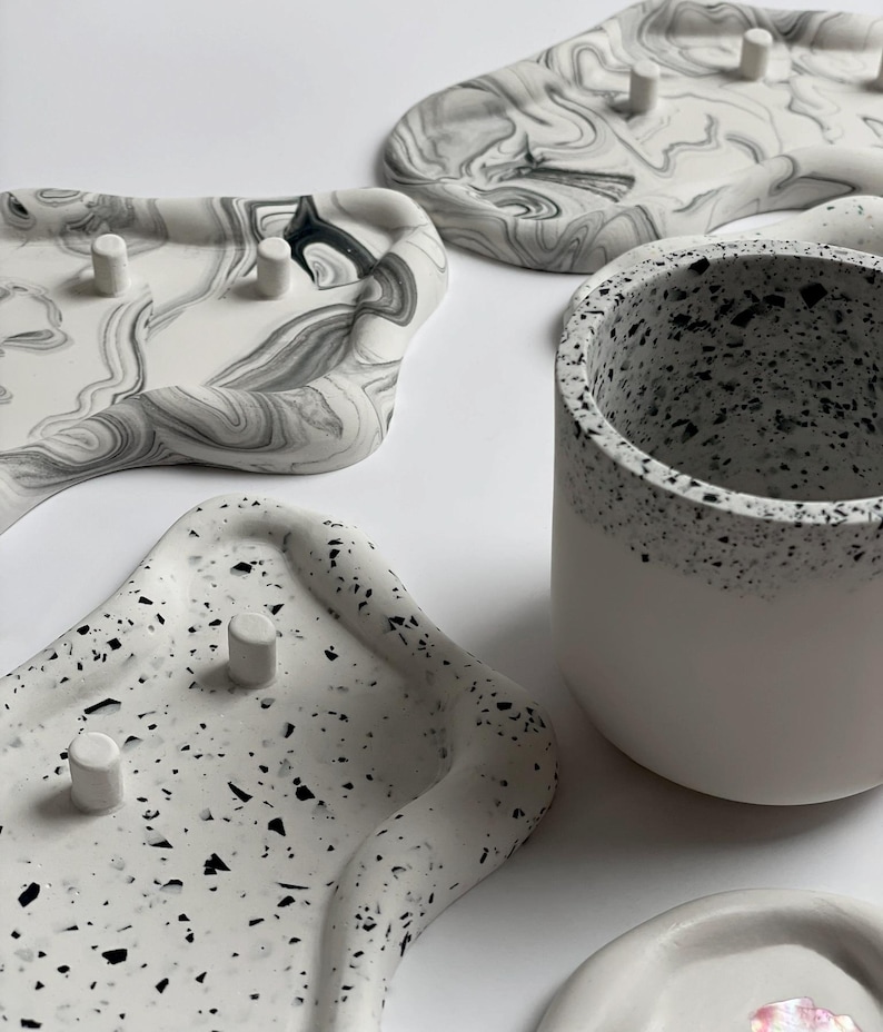 May include: An assortment of decorative trays and a small cup. The trays feature unique designs, including a marbled pattern in shades of gray and white, and a speckled terrazzo-like design. The cup is white with a speckled rim.