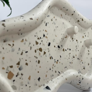 May include: A white, organically shaped planter with a speckled terrazzo design. The planter has a curved rim and two cylindrical feet. The speckled pattern includes black, brown, and tan flecks. The planter is designed for indoor use.