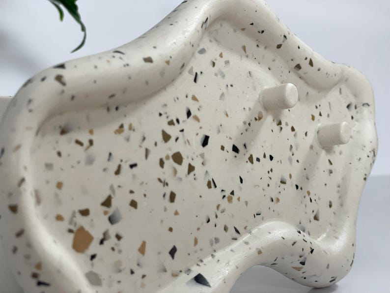 May include: A white, organically shaped planter with a speckled terrazzo design. The planter has a curved rim and two cylindrical feet. The speckled pattern includes black, brown, and tan flecks. The planter is designed for indoor use.