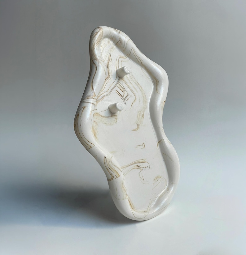 May include: A white, organically shaped tray with a marbled pattern in shades of beige and brown. The tray has two small, cylindrical pegs and a raised edge, suitable for decorative or functional use.