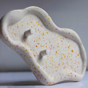 May include: A white soap dish with a speckled design in orange, yellow, and pink. The dish has an organic shape with two cylindrical pegs. The soap dish is a decorative bathroom accessory.