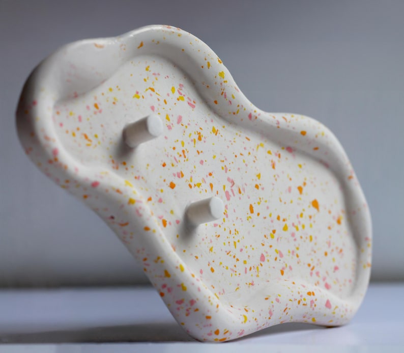 May include: A white soap dish with a speckled design in orange, yellow, and pink. The dish has an organic shape with two cylindrical pegs. The soap dish is a decorative bathroom accessory.