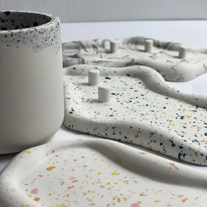May include: A collection of decorative trays and a cylindrical container. The trays are white with colorful terrazzo-style flecks. One tray has small, cylindrical posts. The container is white with a speckled black rim. The items are arranged on a white surface.