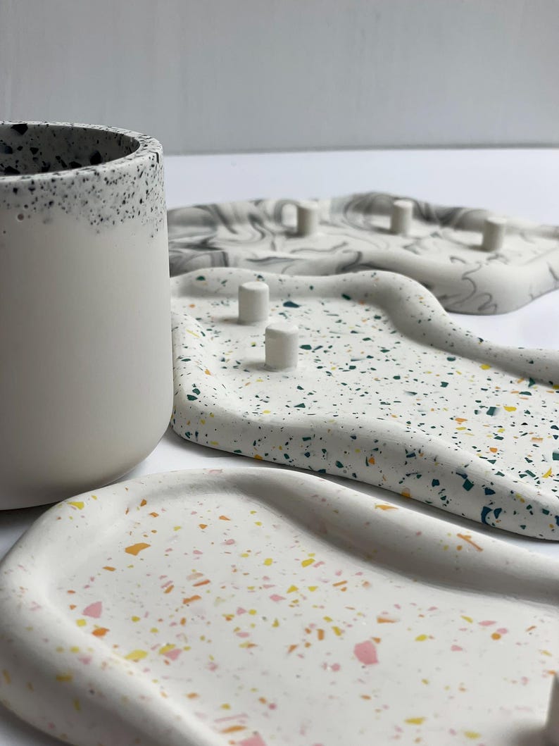 May include: A collection of decorative trays and a cylindrical container. The trays are white with colorful terrazzo-style flecks. One tray has small, cylindrical posts. The container is white with a speckled black rim. The items are arranged on a white surface.