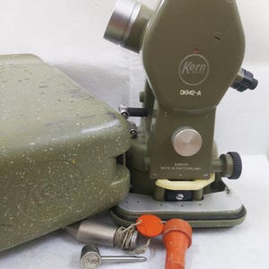 May include: An olive green Kern DKM2-A theodolite with a silver eyepiece and a matching case. The theodolite has a silver base and several adjustment knobs. Included are an orange cap and a silver plumb bob. Made in Switzerland.