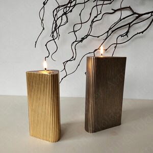 May include: Two lit, rectangular candles with vertical ribbed detailing. One candle is gold, the other is bronze. The candles are on a white surface, with dark, twig-like branches in the background. The candles are lit with a warm glow.