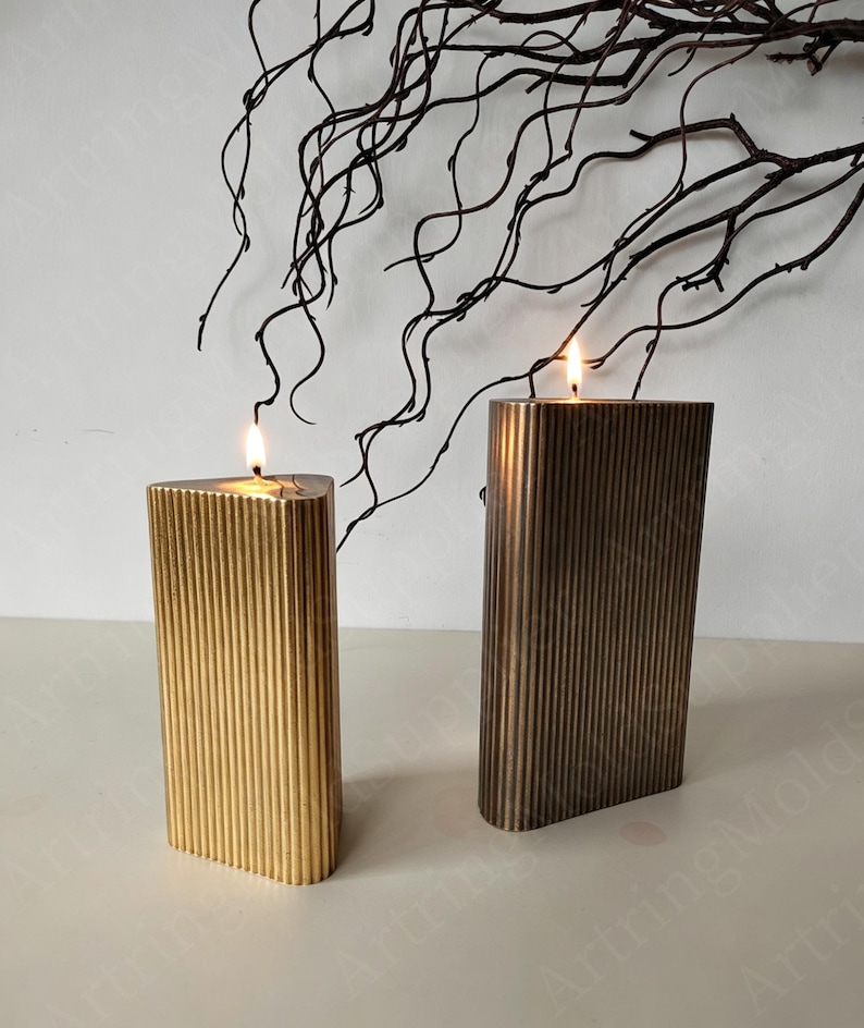 May include: Two lit, rectangular candles with vertical ribbed detailing. One candle is gold, the other is bronze. The candles are on a white surface, with dark, twig-like branches in the background. The candles are lit with a warm glow.