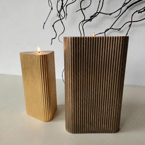May include: Two decorative candles with vertical ribbed designs. One candle is gold and triangular, with a lit wick. The other is bronze and rectangular. Both candles are on a white surface, with dark branches in the background.