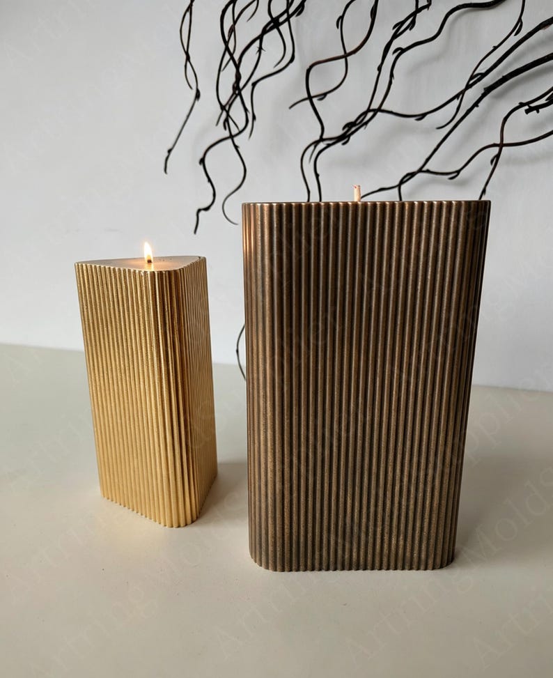 May include: Two decorative candles with vertical ribbed designs. One candle is gold and triangular, with a lit wick. The other is bronze and rectangular. Both candles are on a white surface, with dark branches in the background.