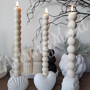 May include: Three white pillar candles with different bases. The candles are decorated with a beaded design. The bases are a seashell, a heart, and a vintage van.