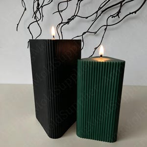 May include: Two lit candles in the image. One is black, the other is dark green. Both candles have a rectangular shape with vertical ribbed detailing. The candles are on a white surface, with dark branches in the background.