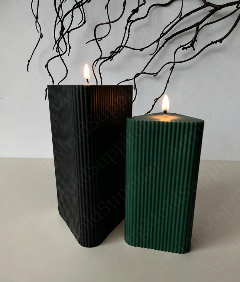 May include: Two lit candles in the image. One is black, the other is dark green. Both candles have a rectangular shape with vertical ribbed detailing. The candles are on a white surface, with dark branches in the background.