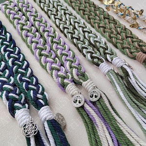 Op de afbeelding: Close-up van gevlochten macramé haaraccessoires in groen, wit, marineblauw en paars. Elke vlecht heeft een decoratieve bedel en hangende strengen. De vlechten worden naast elkaar getoond, waarbij de verschillende kleurencombinaties en ontwerpen worden getoond.