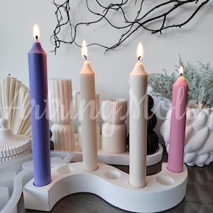 May include: White candle holder with four candle slots, holding three lit candles. The candles are purple, white, and pink. The holder is on a wooden surface.