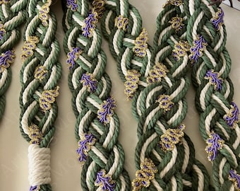Op de afbeelding: Een decoratief gevlochten koord met dikke strengen olijfgroen en crèmekleurig touw die in elkaar zijn gevlochten. Het vlechtwerk is voorzien van kleine, herhalende paarse bloemvormige versieringen omlijnd met glinsterende gouden draad.