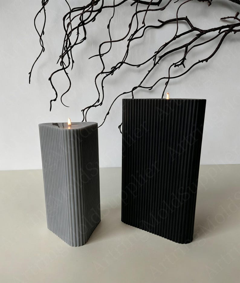 May include: Two lit pillar candles, one gray and one black, with vertical ribbed texture. The candles are rectangular with a slanted top. A dark branch decorates the background.
