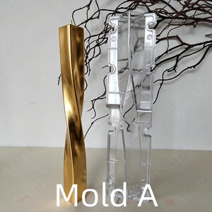 May include: A gold-coloured, twisted candle stands next to a clear plastic mould labelled "Mold A." The candle has a rectangular shape with a spiral design. The mould is designed to create candles. Decorative branches are in the background.