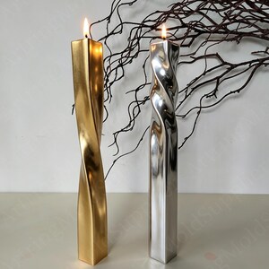 May include: Two lit, twisted pillar candles. One is gold, the other silver. Both have a square base and a spiral design. The candles are on a white surface, with dark brown branches in the background. The flames are bright.