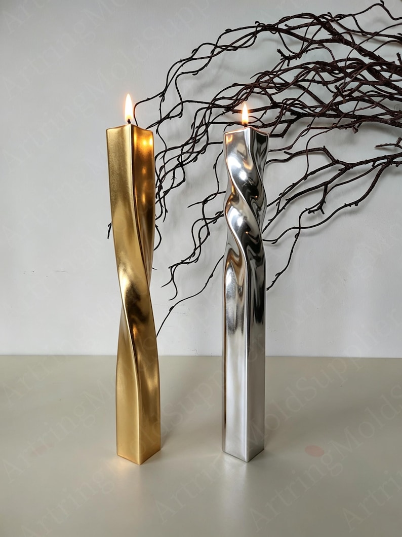 May include: Two lit, twisted pillar candles. One is gold, the other silver. Both have a square base and a spiral design. The candles are on a white surface, with dark brown branches in the background. The flames are bright.