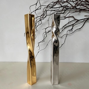 May include: Two tall, twisted candles. One is gold, the other is silver. Both have a square base and a spiral design. The candles are on a white surface, with dark brown branches in the background.