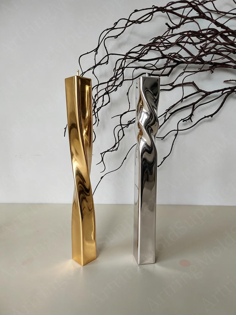 May include: Two tall, twisted candles. One is gold, the other is silver. Both have a square base and a spiral design. The candles are on a white surface, with dark brown branches in the background.