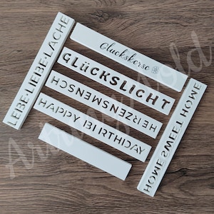 以下が含まれることがあります： 異なるフレーズが切り抜かれた白いプラスチック製のステンシル。フレーズには、「Liebe-Liebe Lache」、「Gluckskersc」、「Glücklicht」、「Hosn」、「He」、「Erzensmen」、「Happy Birthday」、「Howe Meel Howe」が含まれています。