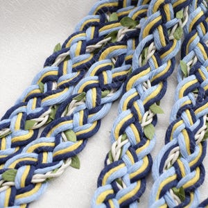 May include: Close-up of braided rope in shades of blue, yellow, and white, with small green leaf accents. The rope is intricately woven, creating a textured pattern. The color palette is bright and cheerful, suggesting a craft or decorative item.