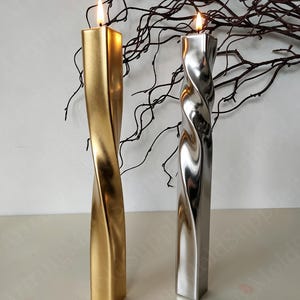 May include: Two twisted, metallic candles with lit flames. One is gold, the other silver. The candles have a square base and a spiraling design. Decorative branches are in the background.