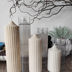 Puede incluir: Cuatro velas blancas en forma de pilar con una textura acanalada, tres de las velas están en recipientes de vidrio transparente con una textura acanalada, los recipientes están sobre una superficie de madera.