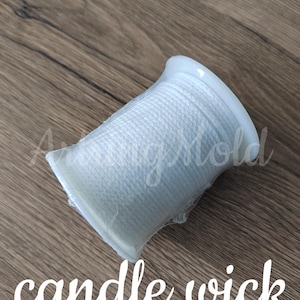 May include: A white candle wick spool, encased in clear plastic, sits on a wooden surface. The words "candle wick" are written in a cursive font beneath the spool. The wick is tightly wound and ready for use.