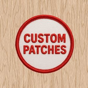 May include: Circular patch with a white center and red embroidered border. The words "CUSTOM PATCHES" are embroidered in red, centered on the white background. The patch is set against a light wood grain background.