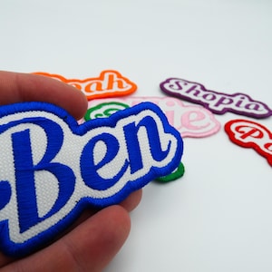 May include: Embroidered name patches in various colours. The patch held in hand says "Ben" in blue script with a white border. Other patches include "Noah", "Shopia", "Poppy", and "Ellie".