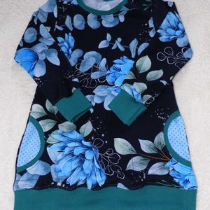 Könnte beinhalten: Langärmliges Kleid mit dunkelblauem Hintergrund und floralem Muster aus großen blauen Blumen und grünen Blättern. Es hat türkisfarbene Bündchen, einen türkisfarbenen Saum und einen türkisfarbenen Ausschnitt. Das Kleid hat zwei Taschen mit hellblauem Polka-Dot-Muster.