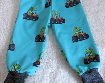 Puede incluir: Pantalones de bebé azules con un patrón de dinosaurio conduciendo un tractor. Los pantalones tienen una cintura y puños elásticos grises.