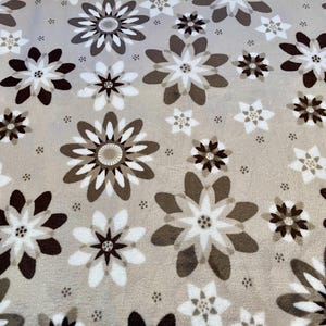 Könnte beinhalten: Ein beigefarbener Stoff mit einem sich wiederholenden Muster aus braunen und weißen stilisierten Blumen. Die Blumen haben ein mehrschichtiges Design mit einem zentralen Kreis.