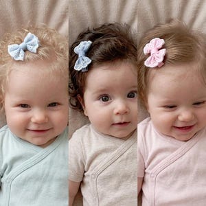 May include: Three babies wearing pastel-colored onesies. Each baby has a different hair color and is wearing a decorative bow hair clip. The bows are light blue and pink, with a delicate, openwork design.