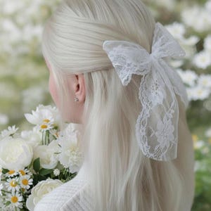Op de afbeelding: Een wit kanten strik haaraccessoire is bevestigd aan de achterkant van het hoofd. Het haar is lang en lichtblond. Een boeket witte bloemen wordt in de armen van de persoon gehouden.