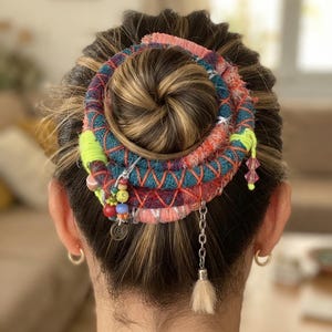 May include: A hair accessory wrapped around a bun, featuring multi-colored yarn in shades of teal, coral, and neon yellow. The accessory is decorated with small colorful beads, a silver-toned metal charm, a dangling chain, and a small beige tassel.