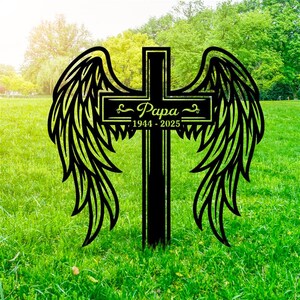 May include: Black metal cross with angel wings, set in a grassy area. The cross features the word "Papa" and the years "1944-2025". The wings are detailed with feather-like cutouts, creating a memorial or decorative piece.