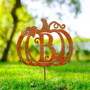 May include: A rust-colored metal garden stake shaped like a pumpkin with a large capital letter B cut out from the center. The pumpkin silhouette features decorative vine and leaf details along the edges and is mounted on a thin metal rod.