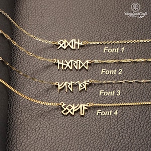 May include: Gold-colored necklaces with pendant designs in four different fonts. The necklaces are displayed on a black leather-like surface. The text "Font 1", "Font 2", "Font 3", and "Font 4" are visible.