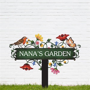 May include: A green metal garden sign with the text "NANA'S GARDEN" in white. The sign is decorated with colorful flowers, a bird, and a butterfly. The sign is mounted on a black post.