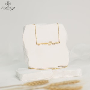 May include: A gold-colored necklace with an arrow design. The necklace is displayed on a white, textured surface. The arrow design features a series of lines and shapes, creating a minimalist and elegant aesthetic. The necklace is delicate and appears to be made of metal.