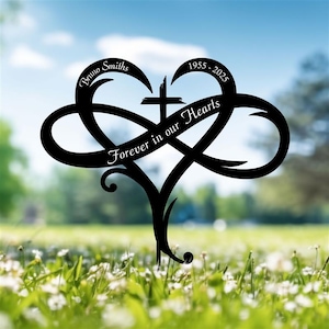 May include: Black metal memorial stake in the shape of a heart intertwined with an infinity symbol. The stake features a cross, the name "Bruno Smiths", dates "1955 - 2025", and the phrase "Forever in our Hearts". The stake is placed in a grassy area.