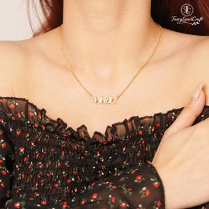 May include: A gold-colored necklace with the word "พอย" in Thai script. The necklace is worn on a person's neck, and the chain is delicate. The person is wearing a black top with a floral pattern.