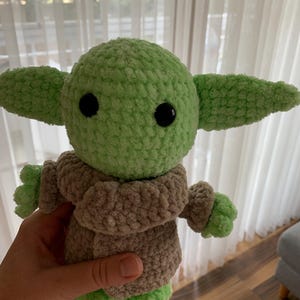 May include: A crocheted plush toy in the likeness of a character from a popular science fiction series. The toy has a light green head and ears, black eyes, and a tan-colored coat. The hands and feet are light green. The toy is approximately 8 inches tall.