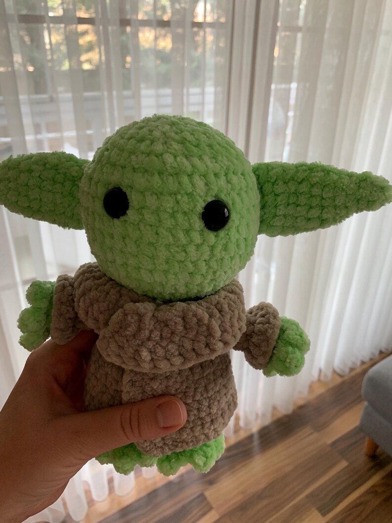 May include: A crocheted plush toy in the likeness of a character from a popular science fiction series. The toy has a light green head and ears, black eyes, and a tan-colored coat. The hands and feet are light green. The toy is approximately 8 inches tall.