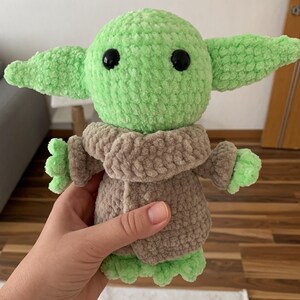 May include: A handmade green and brown crocheted doll, featuring large black eyes and oversized ears. The doll is wearing a brown coat and has green hands and feet. The doll is a character from a popular science fiction franchise.